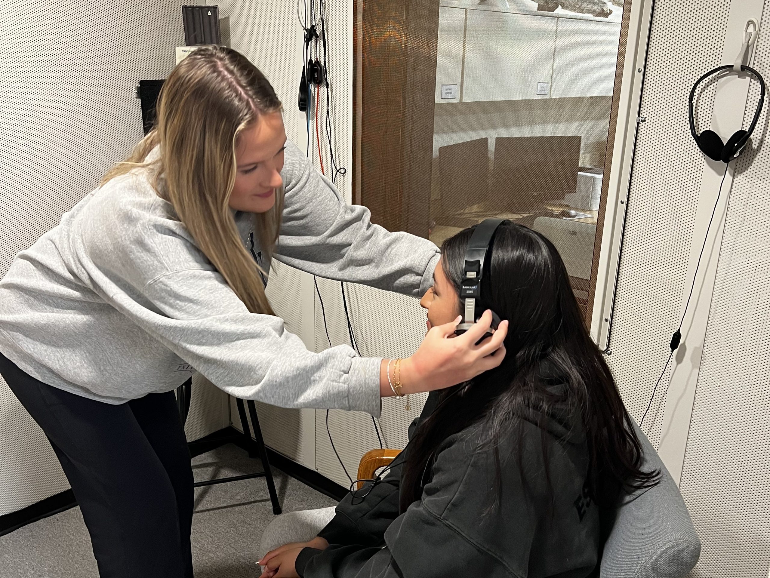 lab2a Students training in the audiology booth. One student puts headphones on another, who is sitting in a chair.